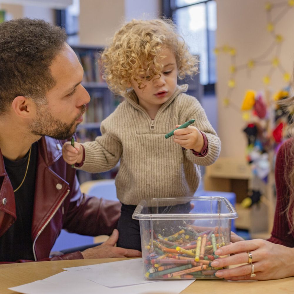 A toddler interacts with two adults while holding a crayon over a container full of crayons in a cozy indoor setting. Cleveland, Ohio, USA