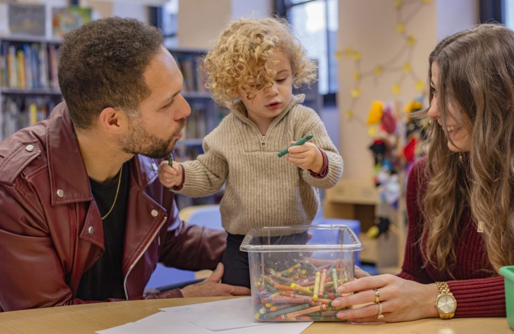 A toddler interacts with two adults while holding a crayon over a container full of crayons in a cozy indoor setting. Cleveland, Ohio, USA